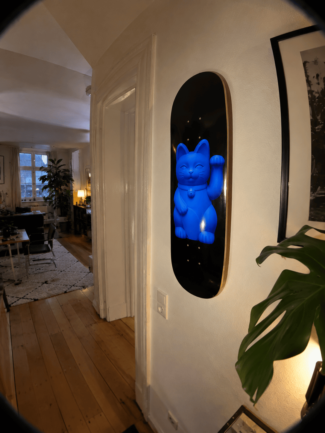 Blue skateboard wall art in a hallway: the Blueprint Lucky Cat deck hung vertically beside a white door frame, monstera leaf in front
