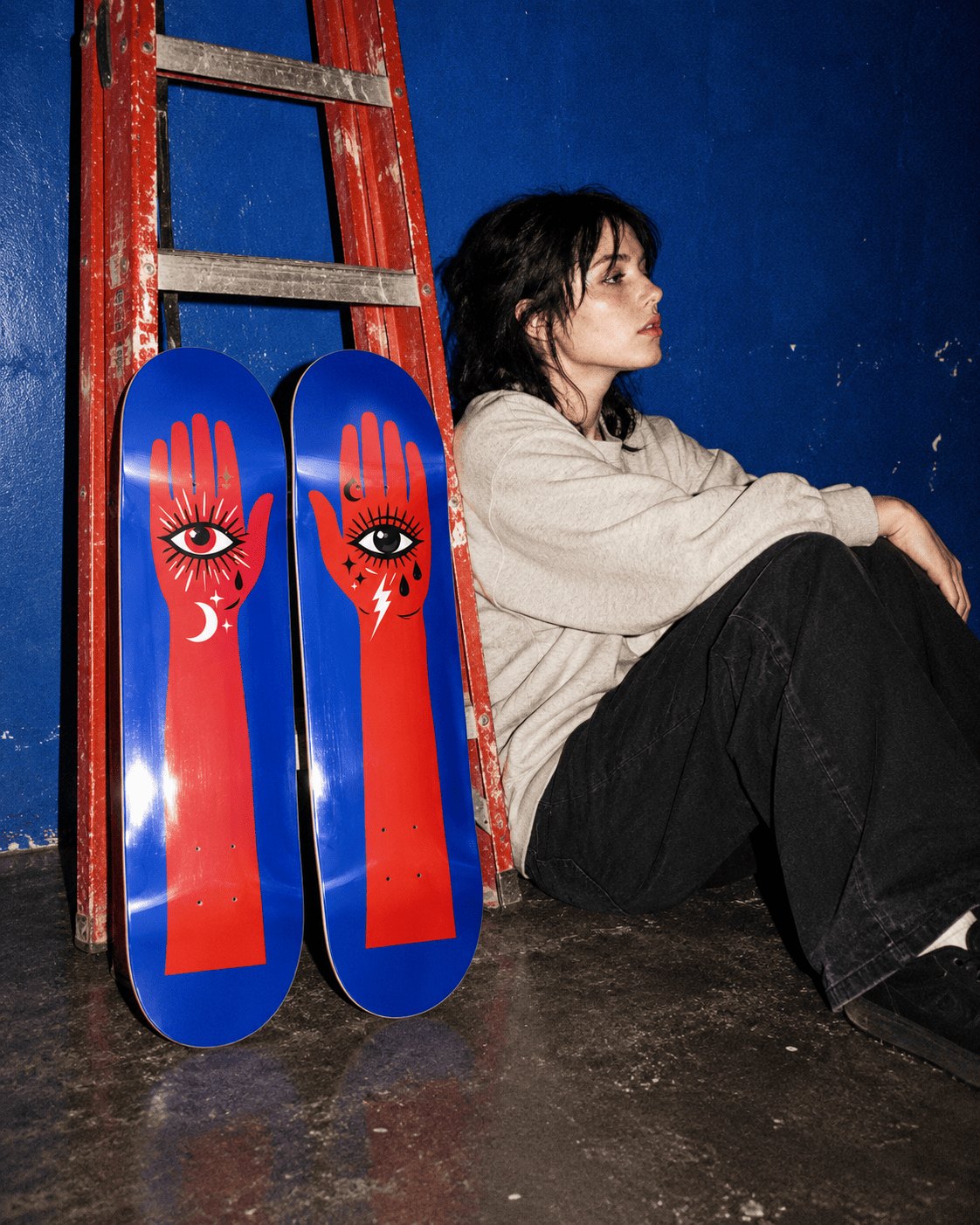 Young woman sitting on a concrete floor next to two blue skateboard decks with a red hand and eye graphic, leaning against a red ladder and a blue wall